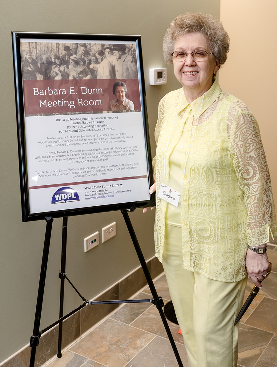 Trustee Barbara E. Dunn smiling next to an info placard if the Meeting Room named after her