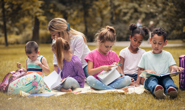 Group of five children with a teacher sitting on a picnic blanket outside with books in their hands
